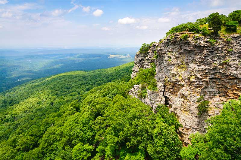 A photograph to represent Arkansas, featuring a view down into a lush green valley full of trees with a large rocky cliffside in the foreground.