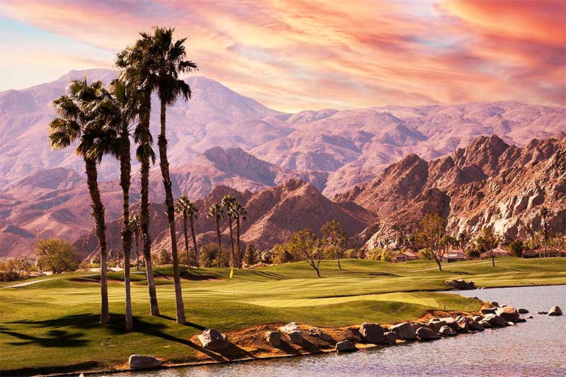 A photograph to represent California featuring palm trees in a large open grassy area alongside a lake. Large mountains rise up in the background, bathed in pink sunset light.