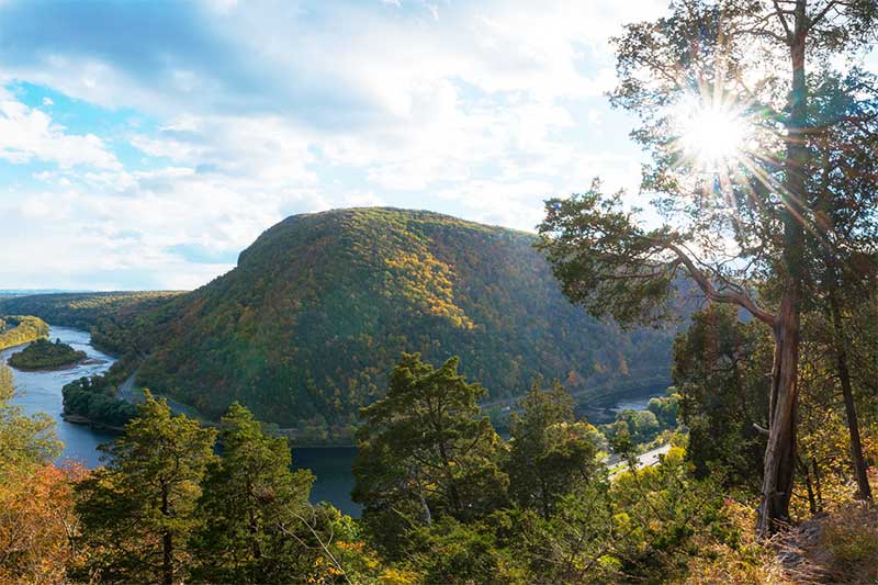 A photograph to represent Delaware, featuring a view from a forested hillside across a river to a large hill covered in trees.