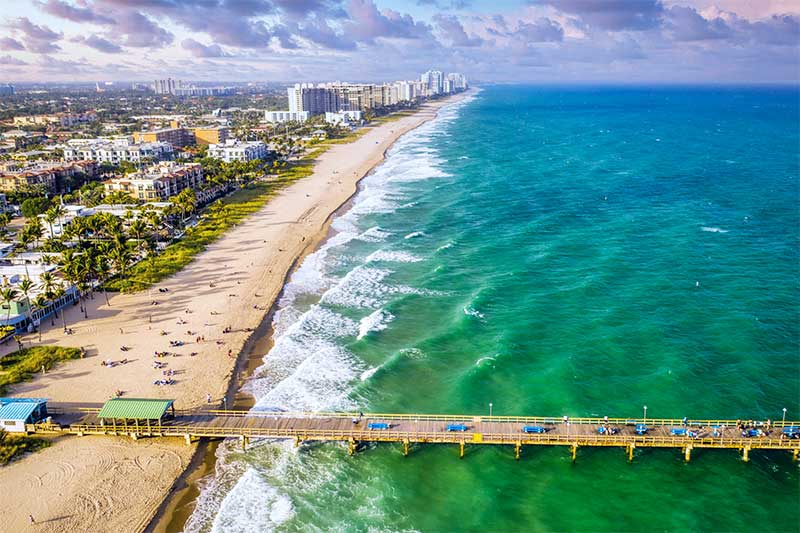 A photograph to represent Florida, featuring an aerial view of a boardwalk extending out into a blue/green ocean from a sandy shoreline lined with palm trees and large resort-like buildings.