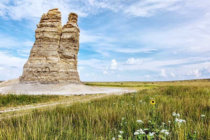 A photograph to represent Kansas, featuring a large rock reaching up toward a blue sky with clouds. It is surrounded by wild grasses.