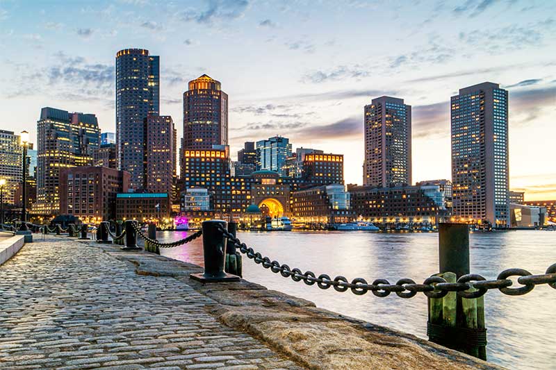 A photograph of the Boston waterfront with iconic skyscrapers in the background. The sun is setting.