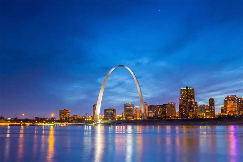 A photograph to represent Missouri, featuring the city skyline and illuminated Gateway Arch at nighttime, viewed from across a body of water.