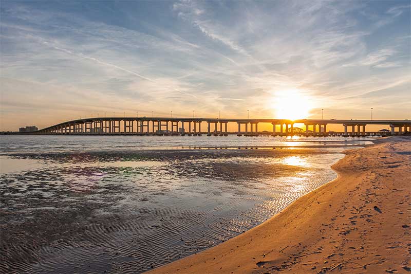 A photograph to represent Mississippi, featuring a sand beach along the water with a large elevated roadway across the water in the background.