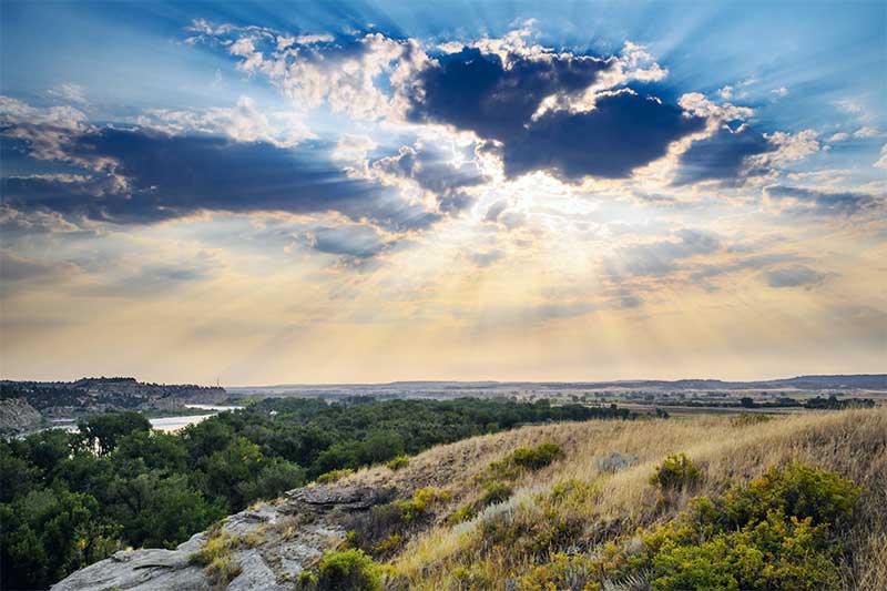 A photograph to represent Montana, featuring a sunburst through fluffy clouds over rocky hillsides blanketed in wild grasses.