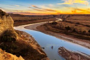 A photograph to represent North Dakota, featuring red and yellow rocky mesa-like structures banking a thin, slow moving river.