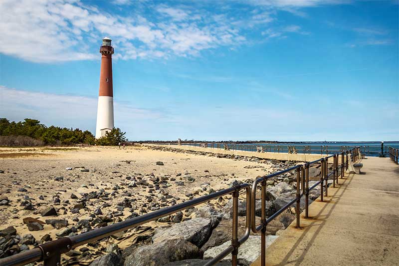 A photograph to represent New Jersey, featuring a lighthouse and cement walkway on a sandy beach dotted with small boulders.