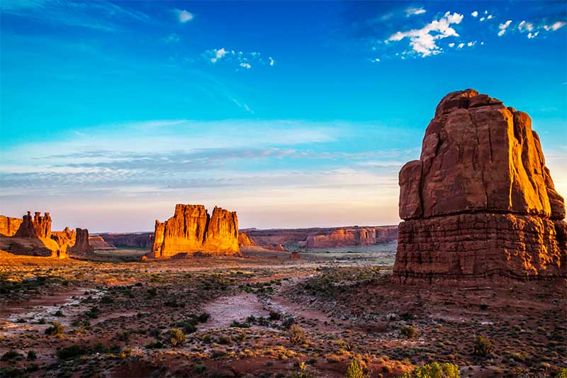 A photograph to represent New Mexico, featuring vivid deep blue skies and flat, rocky landscape dotted with scrubby brush. The occasional immense rock formation emerges from the land, bathed in the golden glow of the setting sun.