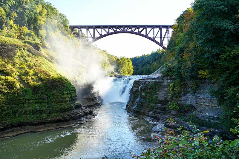 A photograph to represent the state of New York, featuring an arched trestle bridge over a waterfall and river flanked by tall rocky banks.