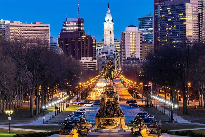 A photograph to represent Philadelphia, featuring a view down an illuminated city street toward the city's skyscrapers.