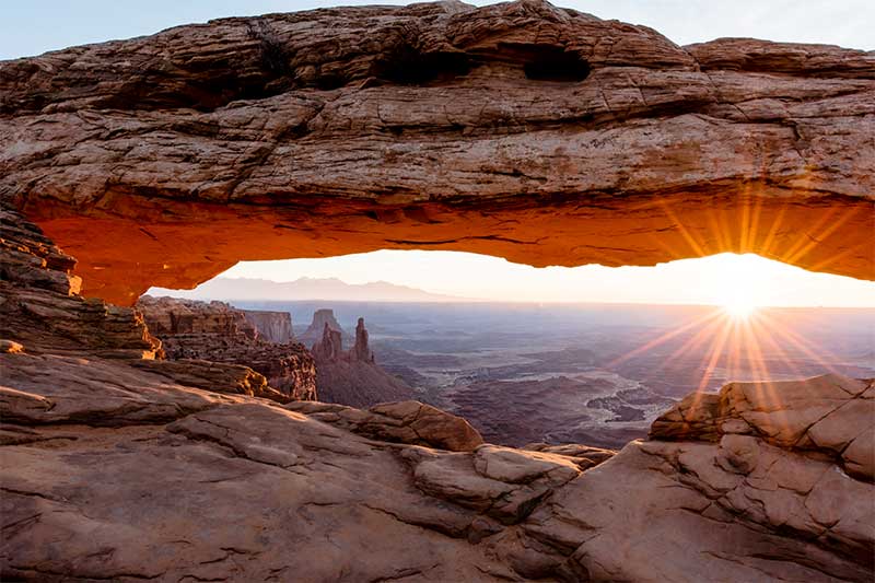 A photograph to represent Utah, featuring the sunset viewed through an opening in a large rock formation on a rocky mountainside overlooking a valley full of canyons.