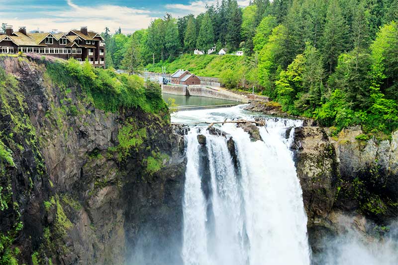 A photograph to represent Washington state, featuring Snoqualmie Falls, crashing down from the top of a rocky landscape covered in bright green trees.