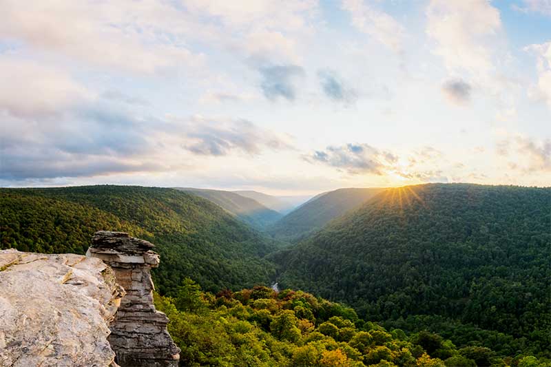 A photograph to represent West Virginia, featuring a rock outcropping and a view from height out across mountainsides covered in green forests.